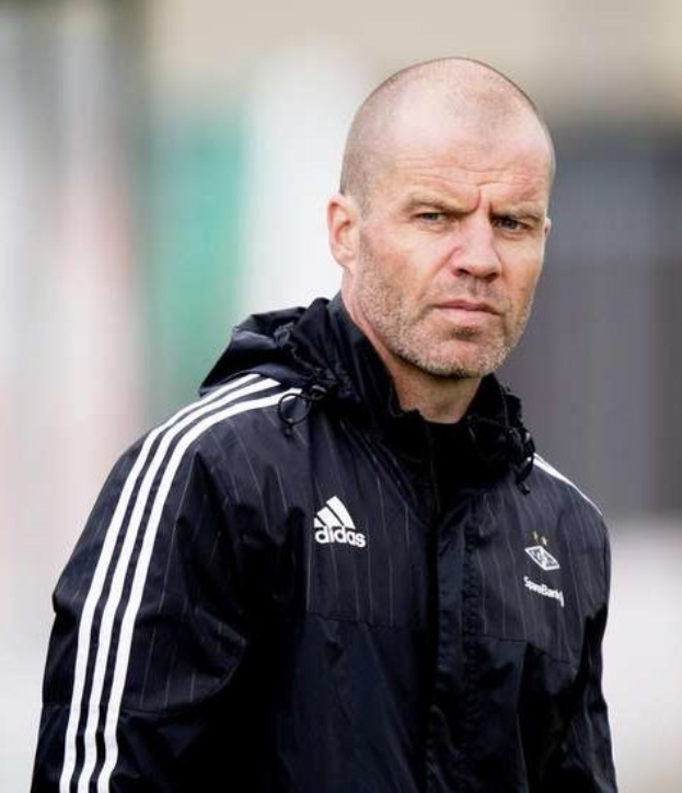 Ein kahlköpfiger Mann in einer schwarzen Jacke, der wie ein Trainer aussieht, steht auf einem Fußballfeld in einer marineblauen Jacke mit weißen Streifen und einem weißen Kragen, die Hände verschränkt und mit einem ernsten Gesichtsausdruck.