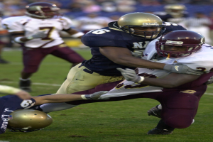 Zwei Fussballspieler in Helmen und Uniformen kämpfen auf dem Feld, während andere im Hintergrund zuschauen.