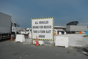 Lastwagen auf einem Parkplatz neben einem Schild mit der Aufschrift "Alle Fahrzeuge nach Mexiko" geparkt mit verstreuten Verkehrskegeln, Brettern und Hintergrundgebäude, Bäume, Pfosten und einem klaren blauen Himmel.