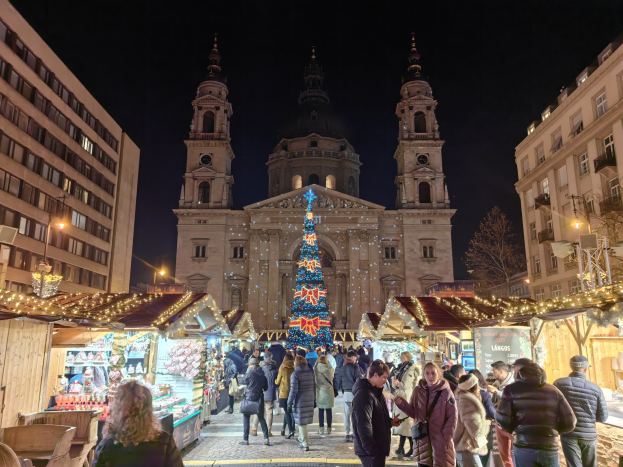 Ein geschäftiger Weihnachtsmarkt mit festlichen Ständen und Lichtern vor einer Kirche bei Nacht, umgeben von Gebäuden, Bäumen und einem sternenklaren Himmel.