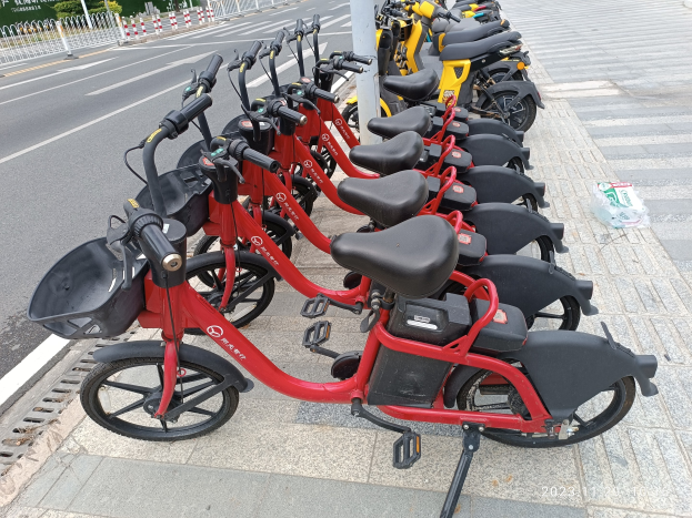Eine Reihe roter und schwarzer E-Bikes, die entlang einer Straße geparkt sind, mit einem weißen Pfosten im Vordergrund und Gebäuden, Bäumen und einem klaren blauen Himmel im Hintergrund.