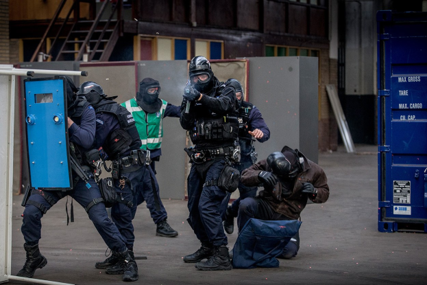 Polizeibeamte in Einsatzausrüstung stehen vor einem Gebäude in einer Reihe, einige halten Waffen, mit einer Person im Vordergrund und Metallobjekten, Treppen und einer Wand im Hintergrund.