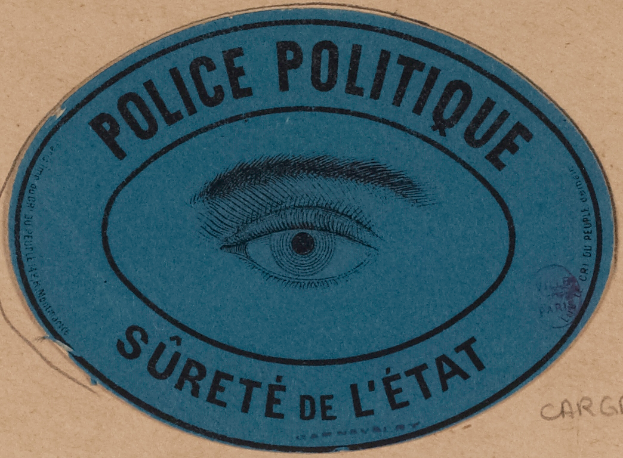 Ein brauner Karton mit einem blauen Aufkleber, auf dem "Polizei Politische Staatssicherheit" steht und ein Auge in der Mitte.