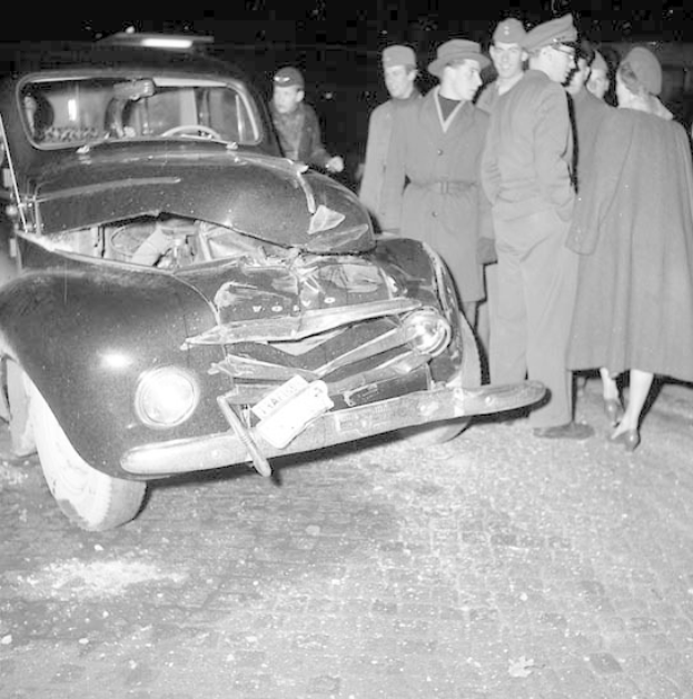 Schwarßlweißes Bild eines schwer beschädigten Autos am Straßenrand mit eingedellter Haube und zersplitterten Fenstern, umgeben von einer Gruppe von Menschen, die die Situation begutachten.
