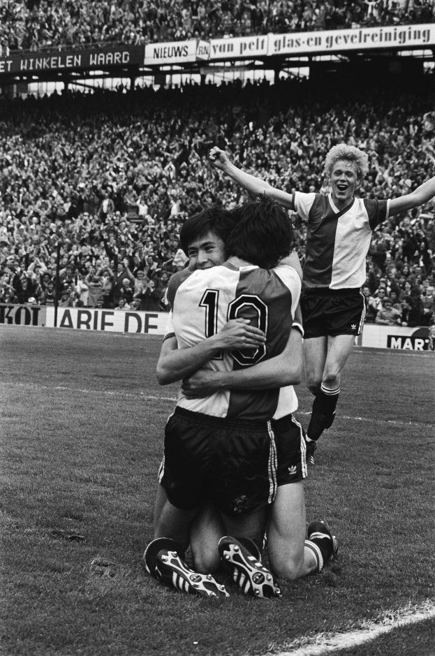 Drei Füßtballer feiern ein Tor, indem sie sich auf dem Boden umarmen, vor einem jubelnden Publikum und schwenkenden Fahnen im Hintergrund, in einem Schwarz-Weiß-Bild.