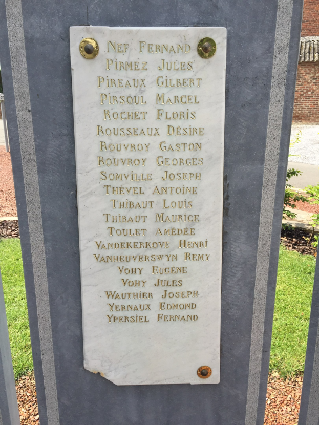Steinbank mit Gedenktafel vor einem Backsteingebäude, umgeben von Bäumen, Pflanzen und Gras.