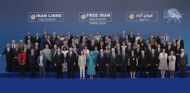 Eine Gruppe von Menschen steht auf einer blauen Bühne mit einem Banner im Hintergrund, auf dem "Free Iran 1000 Ashraf Paris 2018" steht und ein Tierbild zeigt.