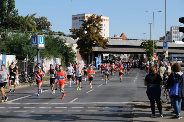 Gruppe von Menschen, die bei einem Marathon auf einer von Bäumen gesäumten Straße laufen, mit Fahrrädern, Schildern, einem Zaun, einer Brücke, Gebäuden und einem klaren blauen Himmel.