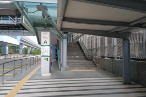 Ein Bahnsteig mit Treppen, eine Straße mit Geländern auf der linken Seite und ein Hintergrund mit Bäumen, Pfählen, Gebäuden und einer Brücke; oben auf dem Bahnsteig befinden sich Rohre und eine Tafel mit Text.