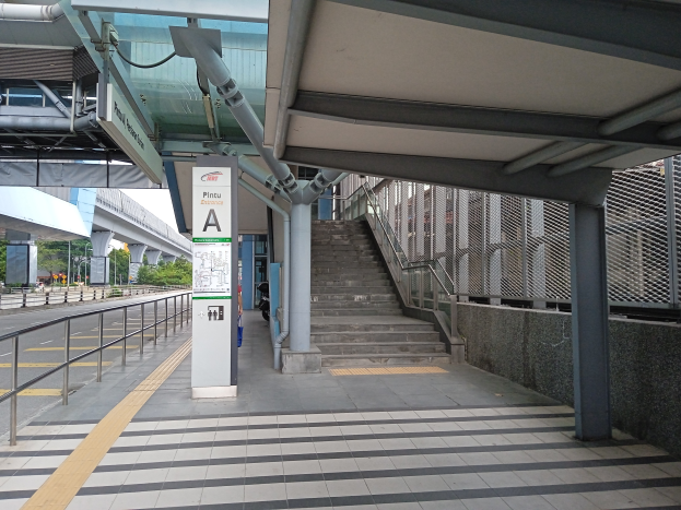 Ein Bahnsteig mit Treppen, eine Straße mit Geländern auf der linken Seite und ein Hintergrund mit Bäumen, Pfählen, Gebäuden und einer Brücke; oben auf dem Bahnsteig befinden sich Rohre und eine Tafel mit Text.
