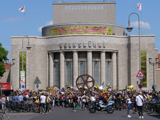 Eine große Gruppe von Menschen versammelte sich vor einem Gebäude mit Säulen und Text, umgeben von Straßenmasten, Straßenlaternen, Schildern, Motorradfahrern, Kraftfahrzeugen, Bäumen und einem bewölktem Himmel, während einer Demonstration in Berlin, Deutschland.