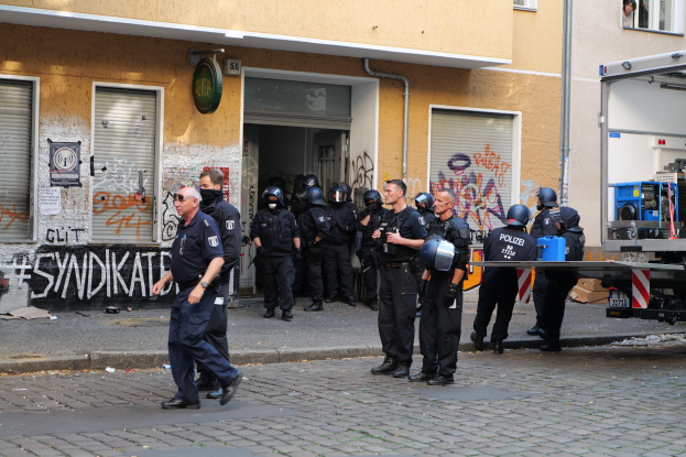 Eine Gruppe von Polizisten, einige mit Helmen und Waffen, steht und geht vor einem Gebäude mit Graffiti, mit einem Fahrzeug und verschiedenen Gegenständen im Hintergrund.