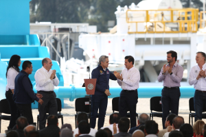 Eine Gruppe von Männern vor einem Publikum mit einem Podium und einem Mikrofon, einige klatschen, Bäume sind im Hintergrund zu sehen.