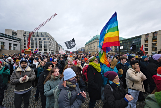 Eine große Gruppe von Menschen auf einer LGBTQ+-Rechtsmarsch in Berlin, die Fahnen und Plakate halten, mit Gebäuden, einem Kran und einem bewölkten Himmel im Hintergrund.
