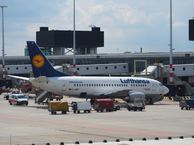 Ein Lufthansa Airbus A320-200 auf dem Frankfurter Flughafen geparkt mit Bodenfahrzeugen und Wagen im Vordergrund, einem Gebäude und Laternen im Hintergrund und einem klaren blauen Himmel.