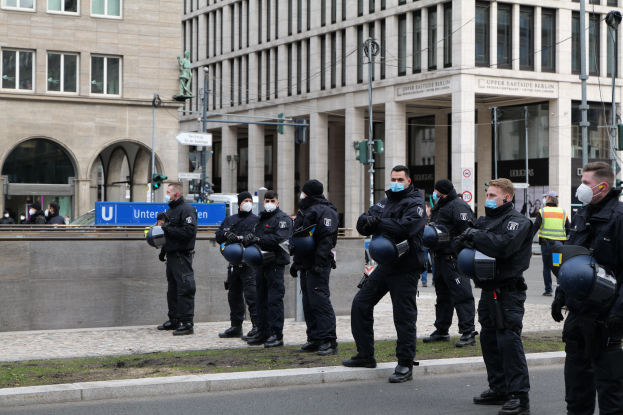 Eine Gruppe von Polizisten in schwarzen Uniformen und Masken steht vor einem Glasfenstergebäude mit Säulen in Berlin, Deutschland, einige halten Helme, mit Laternen, Verkehrsampeln, Schildern mit Text, einer Statue, anderen Menschen und einem grasbewachsenen Boden im Hintergrund.