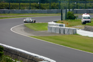 Ein Rennwagen fährt auf einer Rennstrecke mit einem Polizeiwagen im Hintergrund, umgeben von Gras, Pflanzen, Bäumen und Zaun, mit einem Fahrzeug auf der rechten Seite und Text unten.