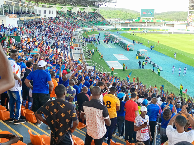 Eine große Menschenmenge steht und sitzt vor einem eingezäunten Fußballfeld, mit Hügeln und einem klaren blauen Himmel im Hintergrund.