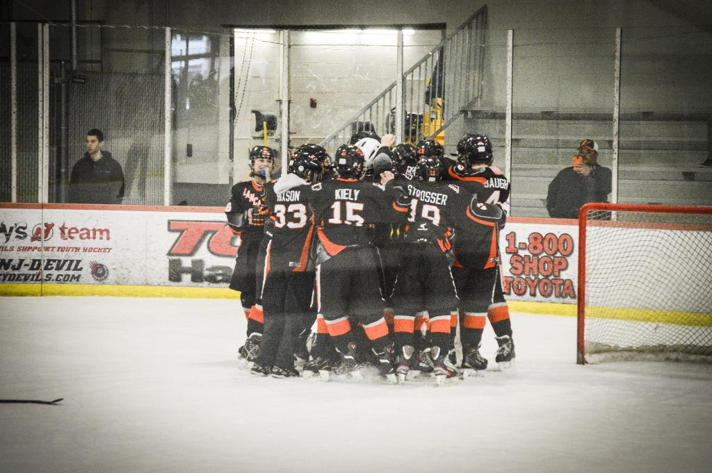 Eine Gruppe von Menschen in Eislaufschuhen, Handschuhen und Helmen steht auf einem Eisgelände mit Absperrungen und Glastüren dahinter, einem Hockey-Tor mit Netz auf der rechten Seite und einem Hockey-Schläger auf der linken Seite.