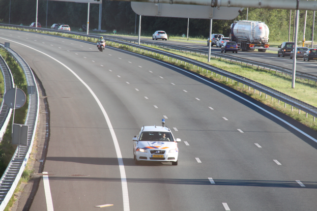 Ein Auto fährt auf einer Autobahn mit einem Polizeiwagen am Rande, anderen Fahrzeugen, Geländern, Pfosten, Planken und Bäumen im Hintergrund.