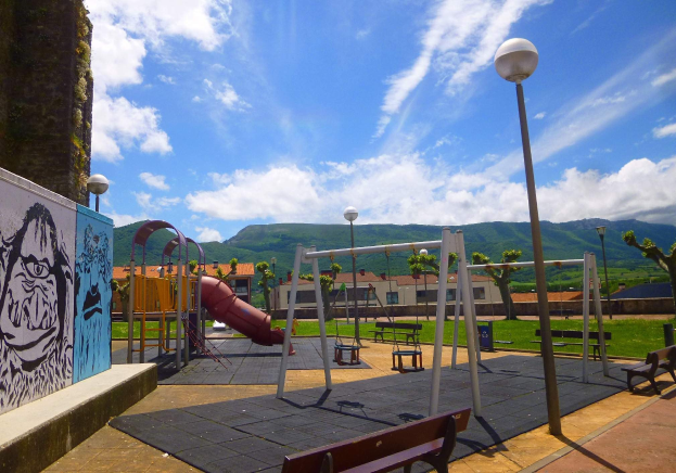 Spielplatz mit Schaukeln, einer Rutsche, Bänken, Masten, Lichtern, einer bemalten Wand, Gras, Bäumen, Gebäuden, Höhen und einem bewölktem Himmel im Hintergrund.