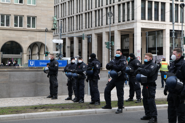 Polizisten in schwarzen Uniformen und Masken stehen vor einem Gebäude mit Glasfenstern und Säulen in Berlin, Deutschland, einige halten Helme, mit Laternen, Verkehrsampeln, Schildern mit Text und einer Statue im Hintergrund.
