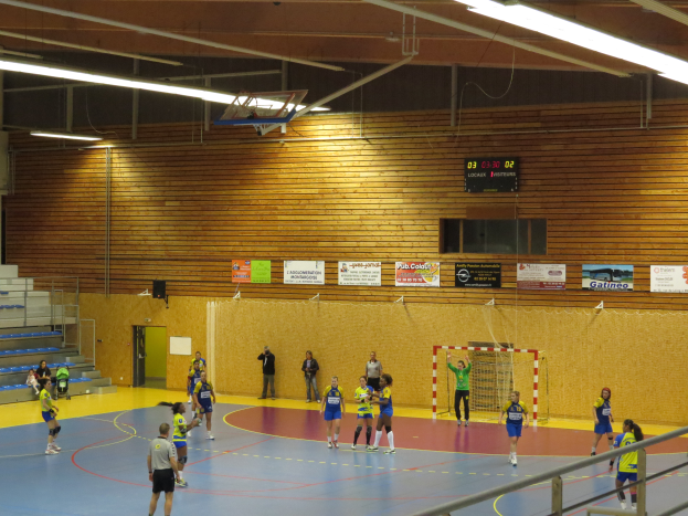 Eine Gruppe von Menschen, die Handball in einer Turnhalle mit sichtbarer Metallzaun, Torstange, Tür, Texttafeln, Anzeigebildschirm, Lautsprecherbox, Metallstangen und Deckenleuchten auf dem Dach mit Metallrahmen spielt.