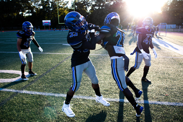 Eine Gruppe junger Männer in Helmen und Fußballschuhen, die auf einem von Bäumen umgebenen Feld mit klarem blauem Himmel Fußball spielen.