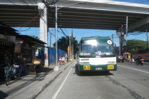 Ein grüner und weißer Bus fährt eine Straße mit Geschäften und Ständen entlang, neben einer Brücke, mit Fußgängern, parkenden Fahrrädern, Strommasten und Bäumen unter einem klaren blauen Himmel.