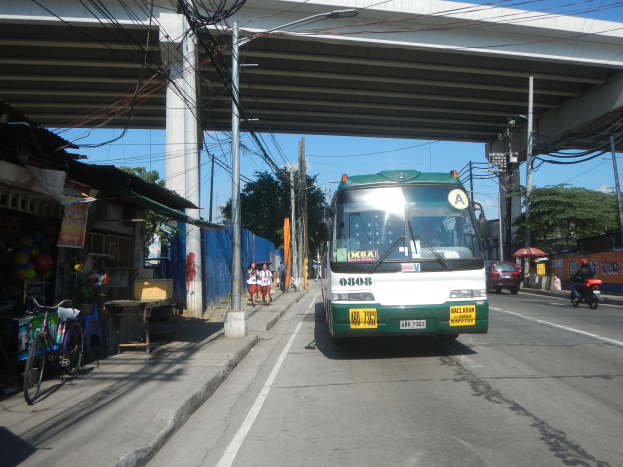Ein grüner und weißer Bus fährt eine Straße mit Geschäften und Ständen entlang, neben einer Brücke, mit Fußgängern, parkenden Fahrrädern, Strommasten und Bäumen unter einem klaren blauen Himmel.