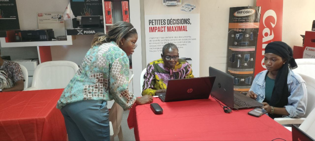 Gruppe von Menschen an einem Tisch mit Laptops und Mobiltelefonen auf einer internationalen Geschäftsmesse in Senegal, mit Bannern, Schränken und einer Uhr im Hintergrund.