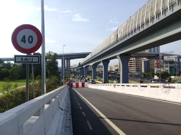 Eine Autobahn mit einem Tempolimit-Schild, Fahrzeuge, eine Brücke mit Pfeilern, Laternen, Bäume, Gebäude und ein bewölkter Himmel.
