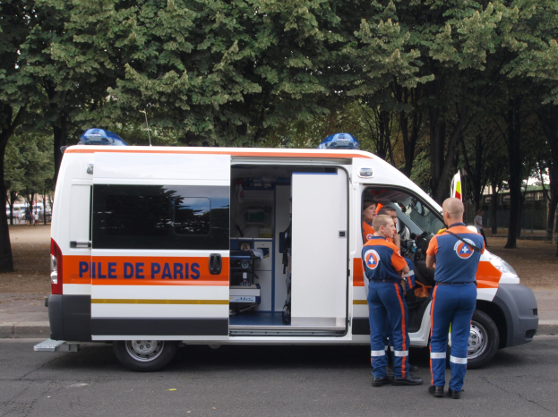 Ambulanz am Straßenrand geparkt mit drei Personen davor, Bäume, Gebäude und andere Fahrzeuge im Hintergrund.