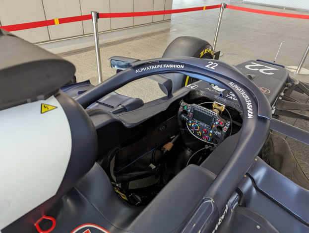 Cockpit eines Formel-E-Autos in einer Ausstellung mit einem Lenkrad im Vordergrund, einer Wand im Hintergrund und dekorativen Bändern um die das Auto umgebenden Pfosten.