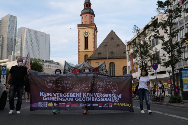 Menschen mit Masken auf der Straße, die eine Fahne halten, mit einem geparkten Auto, Gebäuden, Bäumen, Schildern, Pfosten, einem Uhrenturm und einem klaren blauen Himmel im Hintergrund, was auf eine Protestaktion gegen Rassismus hinweist.