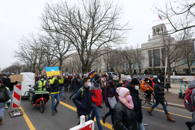 Eine große Gruppe von Menschen marschiert auf einer Straße in Washington, D.C. und hält Schilder, Banner und fährt Fahrräder, mit Bäumen, Schildern und einem klaren blauen Himmel im Hintergrund.