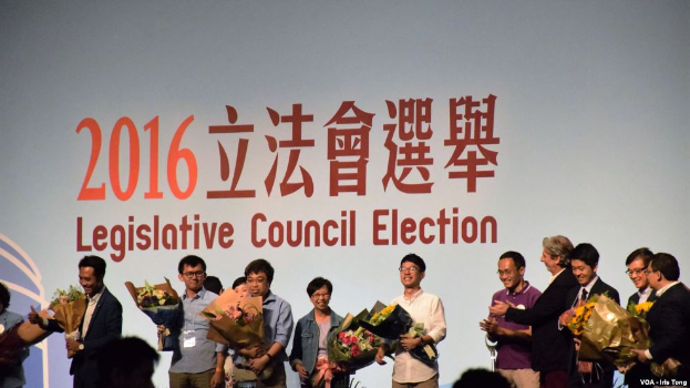 Eine Gruppe von Menschen auf einer Bühne mit Blumensträußen, mit einem Publikum darunter und einem Bildschirm im Hintergrund mit Text und einem Logo für die Wahl des Legislativrats 2016.