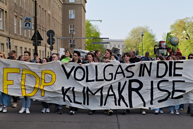 Eine Gruppe von Menschen marschiert mit einem Banner, auf dem "FDP Vollgas in die Klimakrise" steht, eine Straße entlang, mit Gebäuden, Bäumen und Verkehrszeichen im Hintergrund unter einem klaren blauen Himmel.