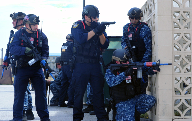 Gruppe von Polizisten in Helmen und Uniformen auf einer Brücke stehend, Waffen haltend, mit einer Wand rechts und Gebäuden, Pfählen, Bäumen und Himmel im Hintergrund.