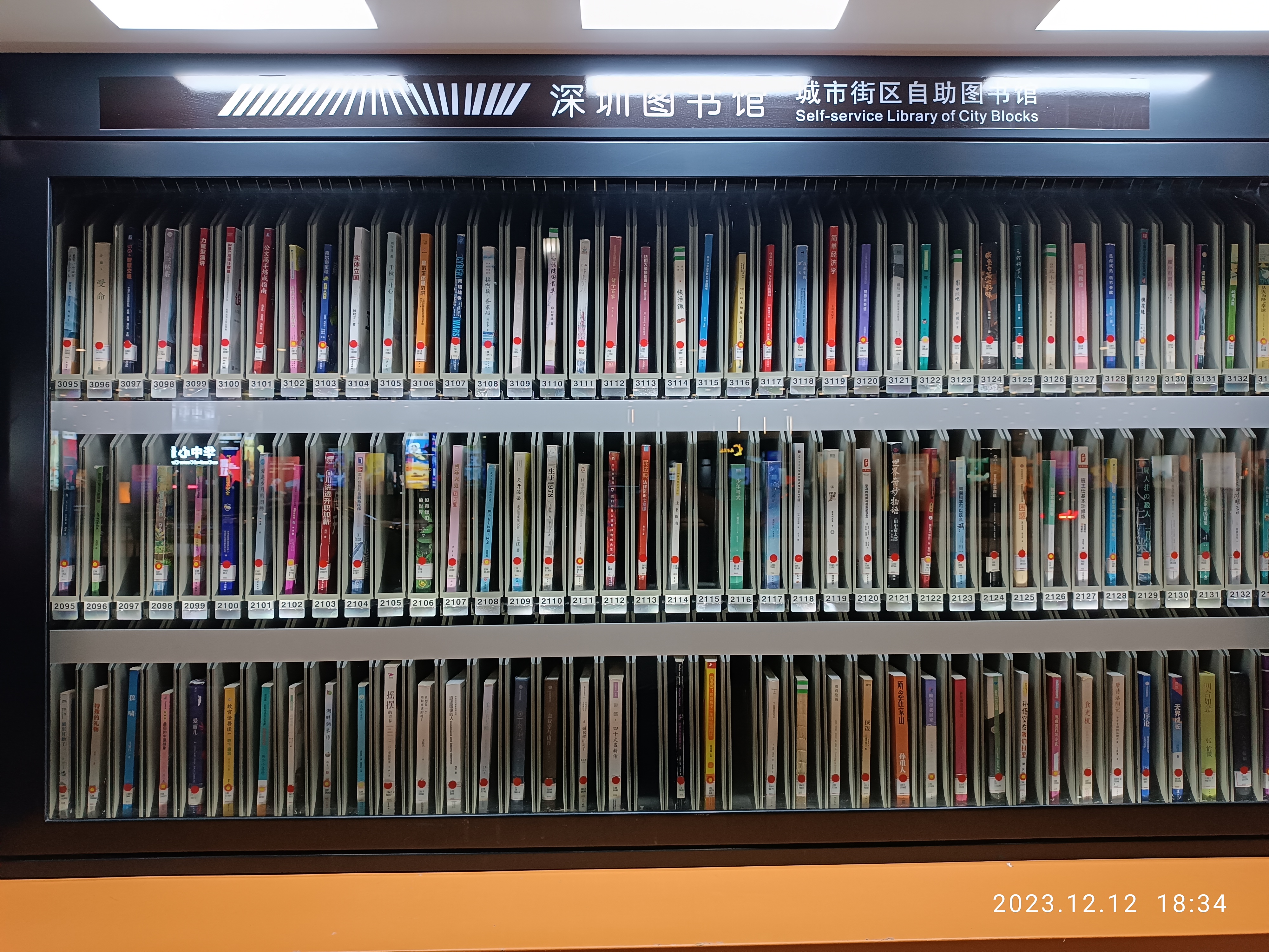 Eine Bibliothek mit zahlreichen Büchern auf Regalen, beleuchtet von Deckenlampen, mit einer Texttafel und einem Wasserzeichen unten, ähnlich einer Selbstbedienungsbibliothek von Stadtblöcken.