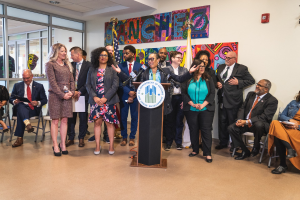 Gruppe von Menschen vor einem Rednerpult mit Mikrofon und einer Tafel mit dem Text "Sancho City Councilman Joins City Council" steht, mit sitzenden und stehenden Gästen, Flaggen, Feuerlöscher und Deckenleuchten im Hintergrund.