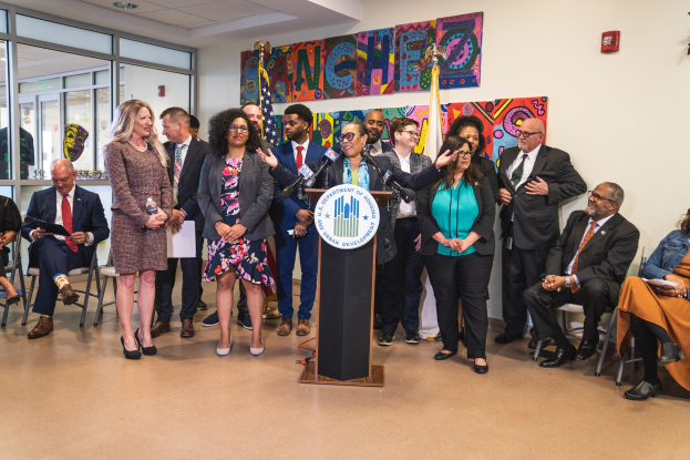 Gruppe von Menschen vor einem Rednerpult mit Mikrofon und einer Tafel mit dem Text "Sancho City Councilman Joins City Council" steht, mit sitzenden und stehenden Gästen, Flaggen, Feuerlöscher und Deckenleuchten im Hintergrund.