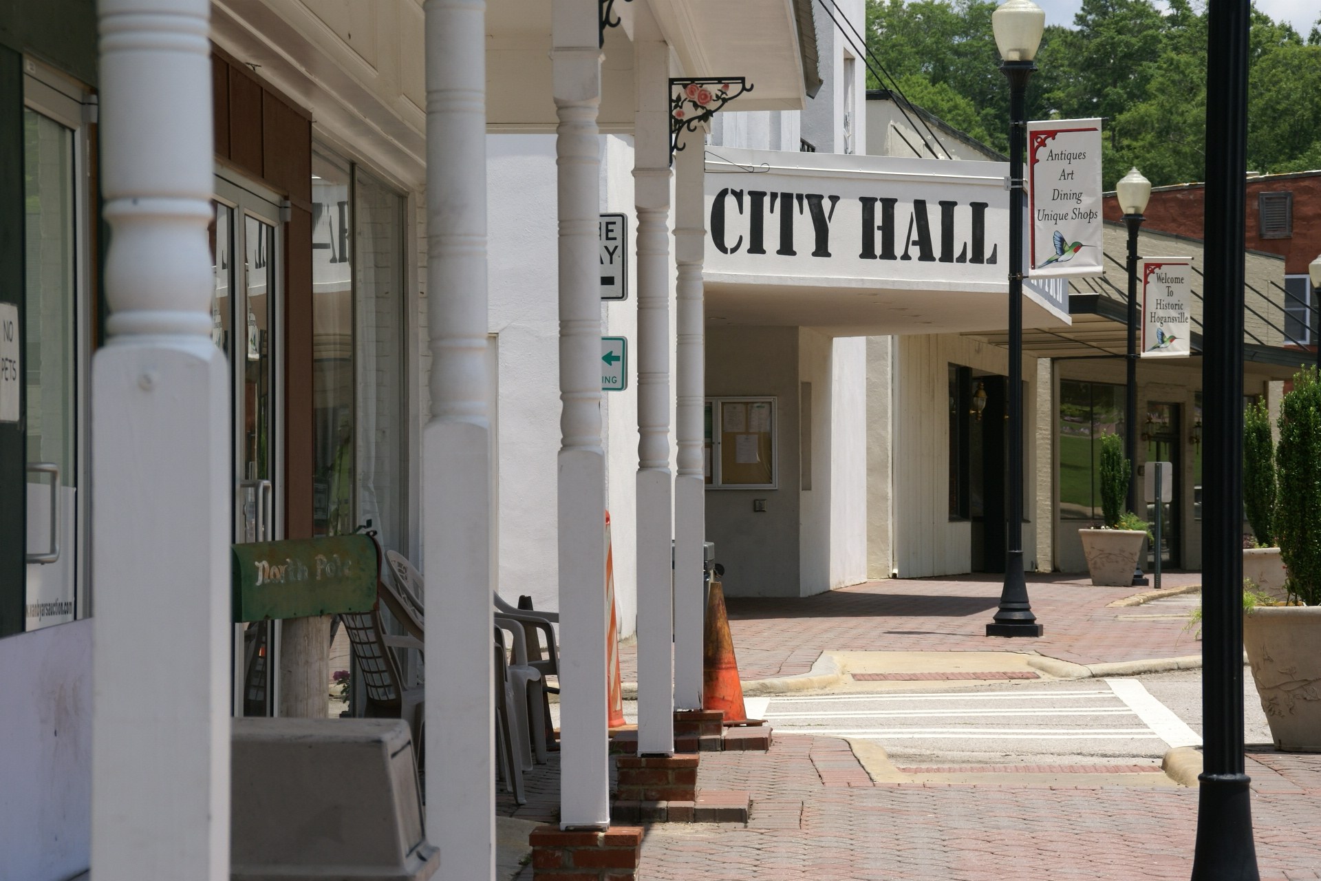Ein Rathausgebäude mit umliegender Infrastruktur, darunter Straßenmasten, Leuchten, Namenschilder, Fahrzeuge, Verkehrskegel, Stühle, Topfpflanzen, Bäume und ein bewölkter Himmel.