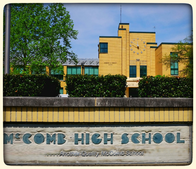 Eine Wand mit der Aufschrift "MCCOMB HIGH SCHOOL", mit Pflanzen, Bäumen, einem Pfahl, einem Gebäude und einem bewölkten Himmel im Hintergrund.
