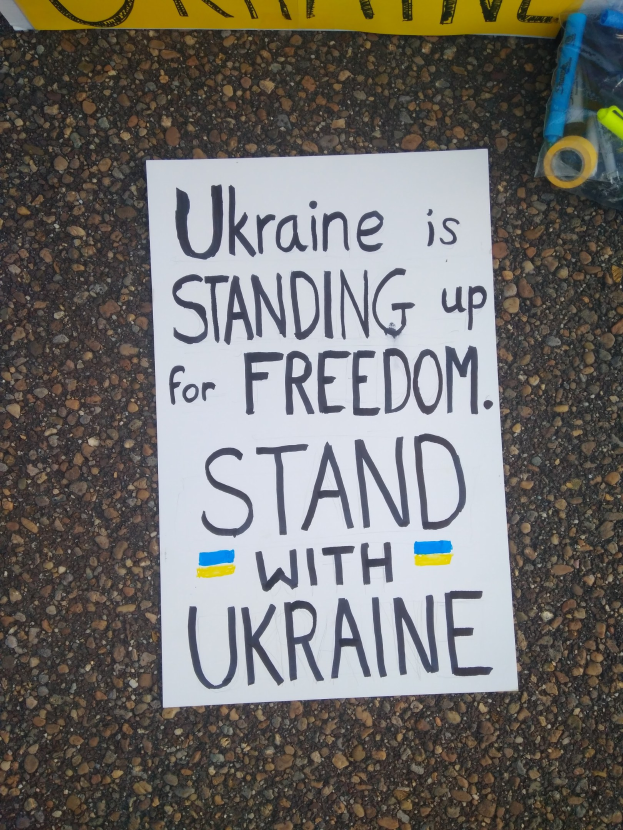 Ein weißes Blatt Papier mit dem Text "Ukraine steht für Freiheit ein, steht mit Ukraine" vor einer gelben Fahne auf dem Boden. Rechts davon ein Plastikbehälter mit verschiedenen Gegenständen.