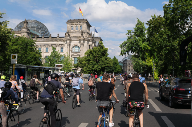 Eine Gruppe von Menschen, die auf Fahrrädern eine von Bäumen gesäumte Straße in Berlin, Deutschland, entlangfahren, mit Gebäuden, einem Bushaltestellenschild auf der rechten Seite und einer Flagge, die auf einem der Gebäude weht, unter einem bewölkten Himmel.