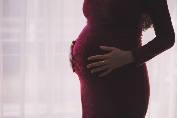 Eine schwangere Frau in einem roten Kleid steht vor einem Fenster, Hände auf dem Bauch, mit einem sanft unscharfen Hintergrund.