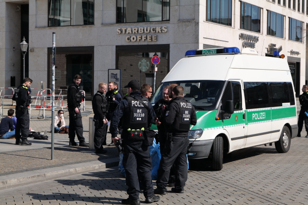 Eine Gruppe von Polizisten steht vor einem Starbucks-Café, rechts ein Van und links ein paar Menschen, im Hintergrund ein Gebäude mit Fenstern, einem Schild, einem Laternenmast und einem Zaun.