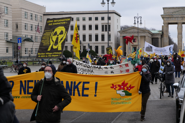 Eine große Gruppe von Menschen marschiert mit Schildern und Fahnen auf einer Straße während einer Demonstration gegen Atomkraft in Deutschland, mit Fahrzeugen auf der rechten Seite und Gebäuden, Laternen, Bäumen und einem Tor im Hintergrund.