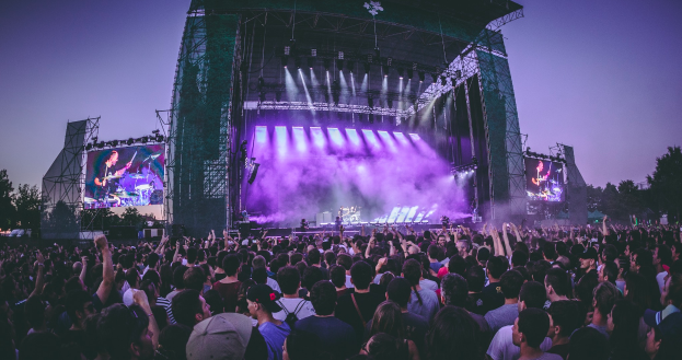 Eine große Menge Menschen auf einem Musikfestival, mit einer Bühne in der Mitte, auf der Musiker Instrumente spielen, beleuchtet von Lichtern und umgeben von Bildschirmen, und einem Gebäude, Bäumen und einem klaren blauen Himmel im Hintergrund.